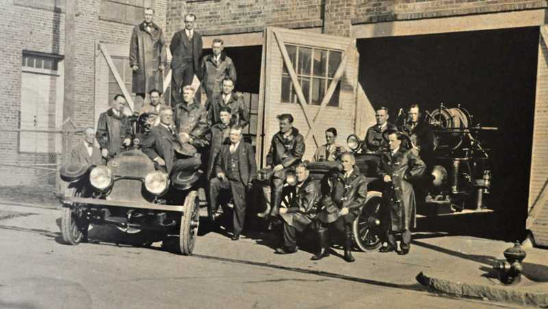Black and white photo of firefighters posing around two antique firetrucks. 